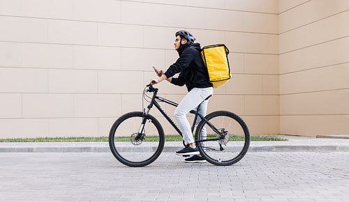 Side view of a young messenger with termal backpack man riding his bike and using mobile phone in the city