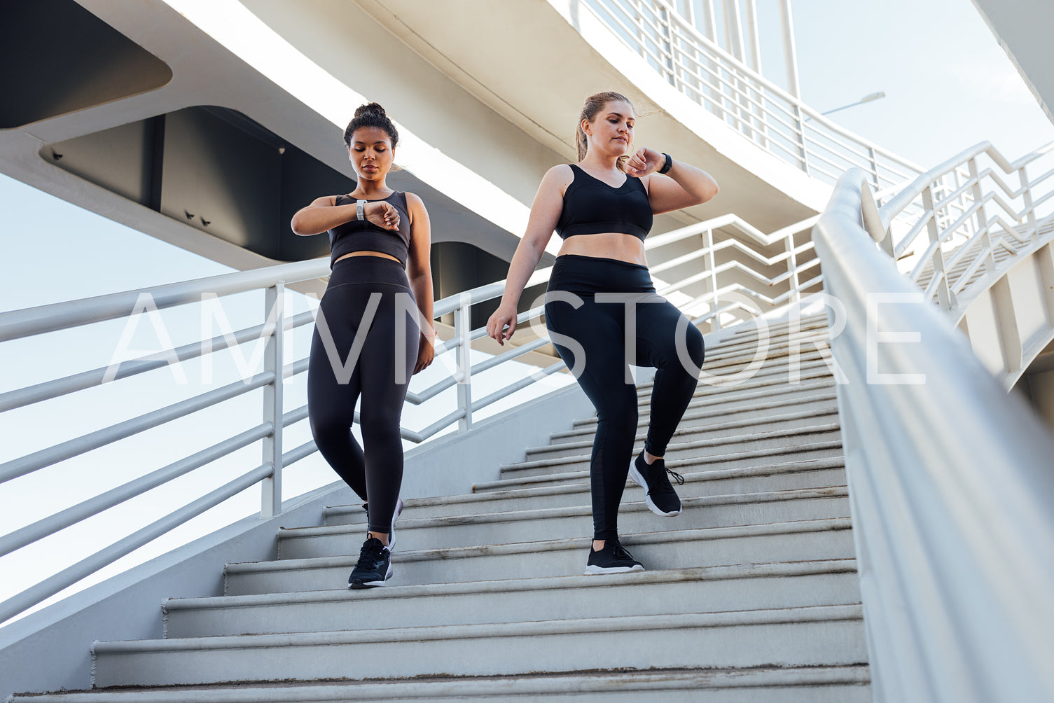 Two young females go on the stairs and watch on their smartwatches. Plus-sized woman with her friend looking at smartwatch while stepping down on stairs.