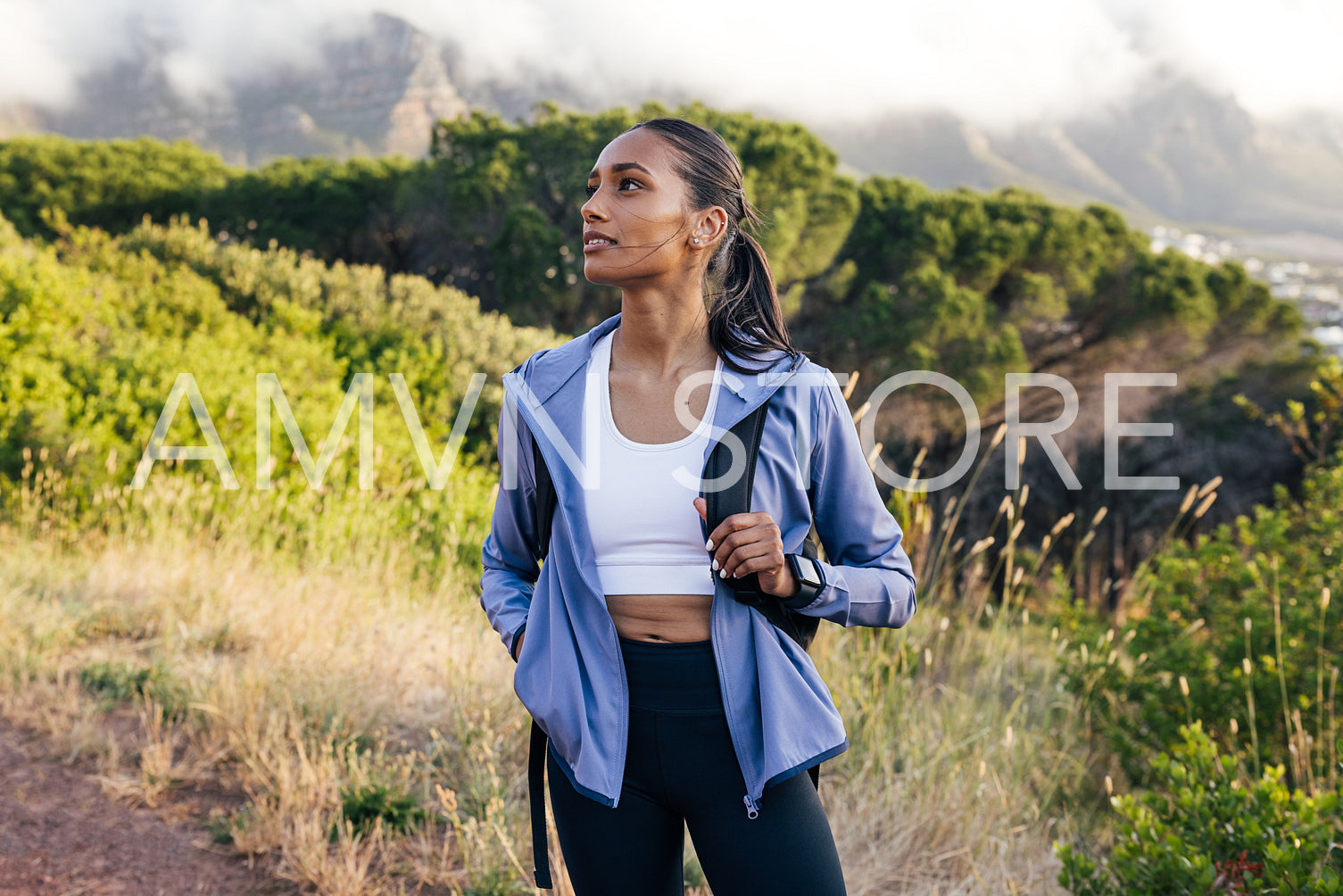 Woman hiker in sportswear looking away while standing against mountain with clouds at sunset. Female trail runner taking a break. 