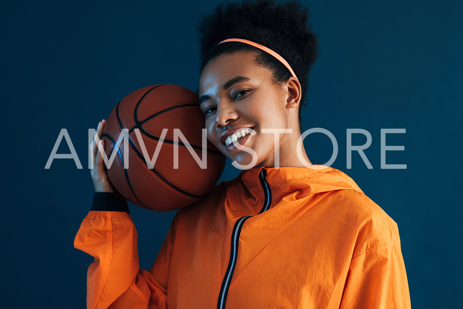 Portrait of smiling female with basketball on her shoulder looking at camera. Cheerful woman in orange fitness attire posing with a basketball over a blue backdrop.