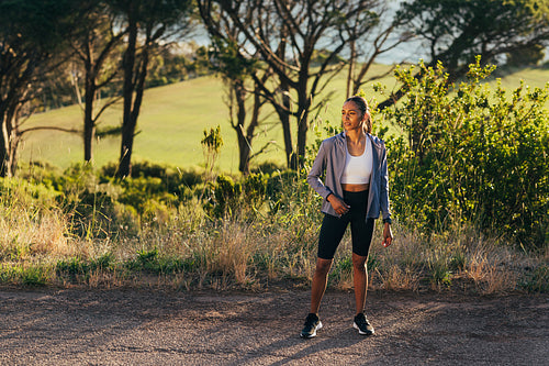 Full length of slim female in sportswear standing outdoors at sunset, taking a break