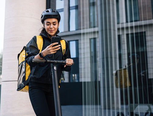 Smiling delivery woman with backpack looking on smartphone leaning on a handlebar of electric push scooter