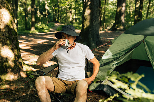 Senior tourist drinks coffee while sitting on chair in the forest near his tent