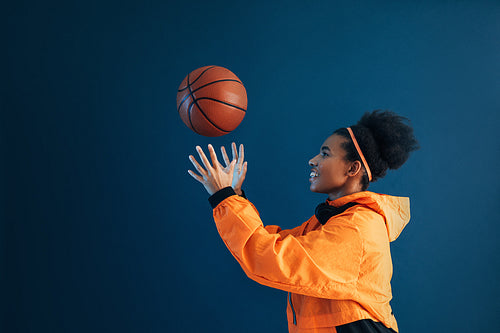 Side view of smiling woman in orange fitness attire catching basketball over blue backdrop