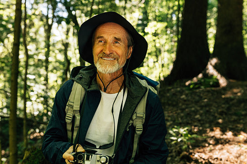 Cheerful senior male with a backpack and film camera taking a break during a forest walk