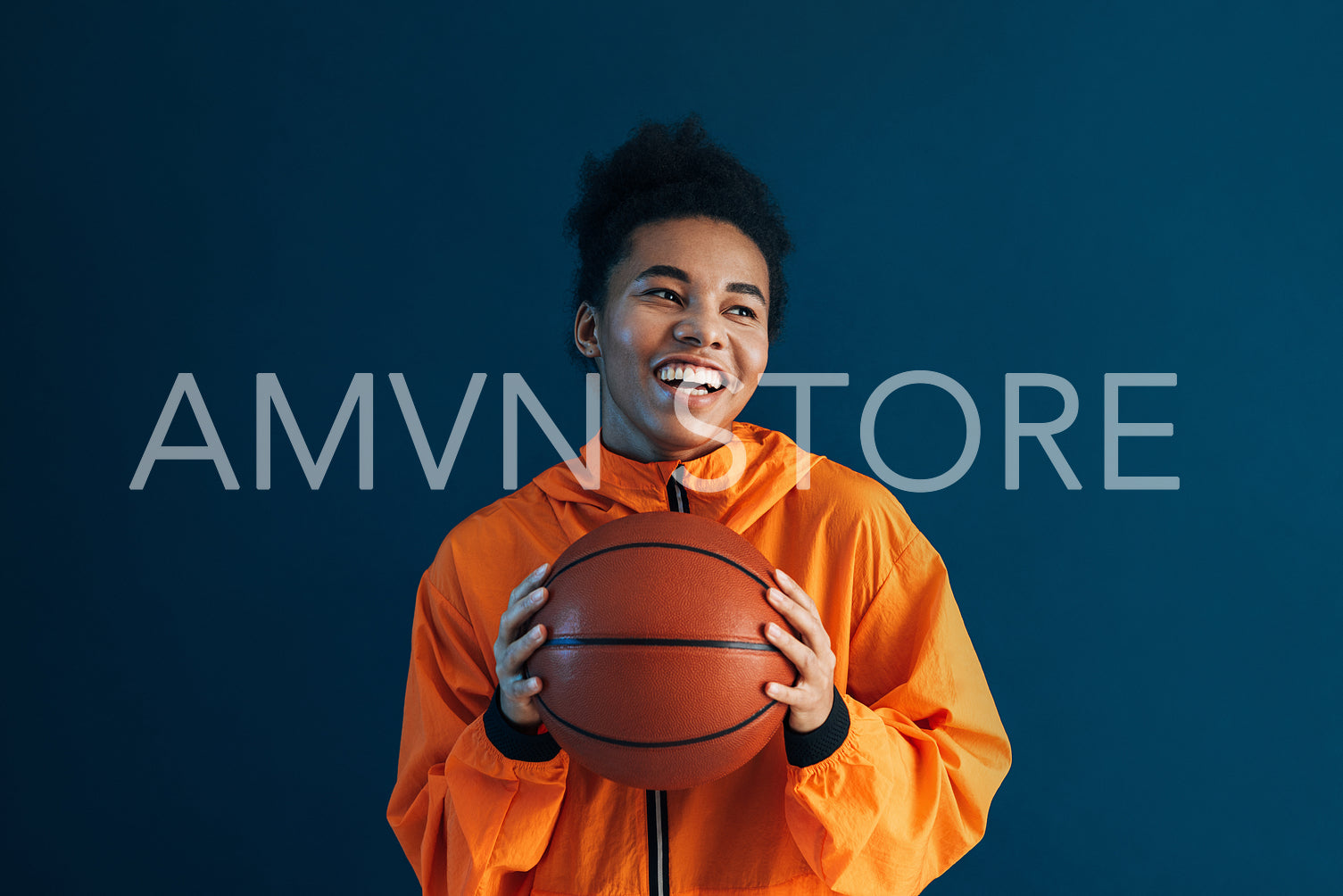 Portrait of a happy woman in orange sportswear holding basketball. Young cheerful female over a blue backdrop with basketball.