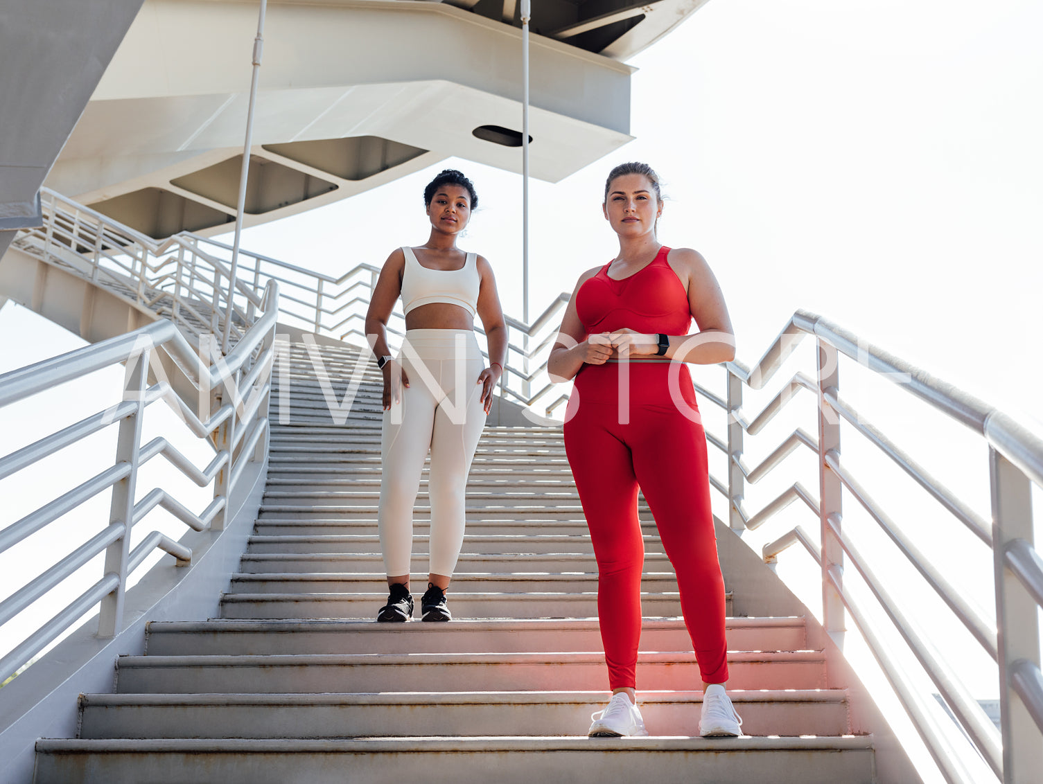 Two females in fitness attire with different colors looking at camera while standing on a staircase. Plus-sized woman with her fitness friend posing together.