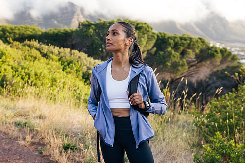 Woman hiker in sportswear looking away while standing against mountain with clouds at sunset. Female trail runner taking a break.