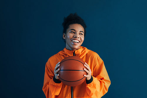 Portrait of a happy woman in orange sportswear holding basketball. Young cheerful female over a blue backdrop with basketball.