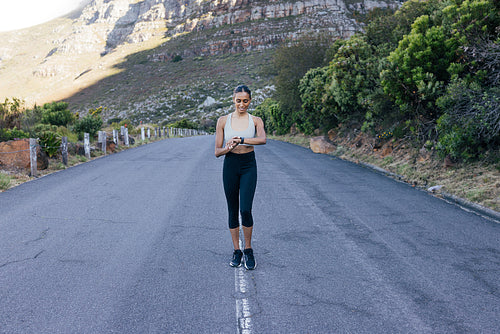 Full length of a smiling female walking on an abandoned road and checking her pulse. Cheerful woman using smartwatches while doing morning walk.