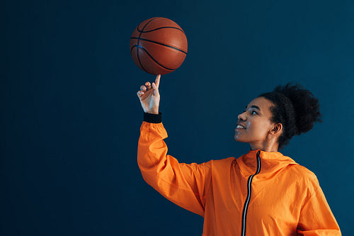 Professional female basketball player spins a basketball on finger over the blue backdrop. Young woman in orange fitness attire in studio.