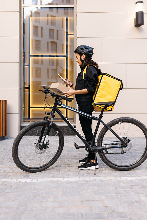 Side view of woman courier with backpack and bicycle standing at apartment building holding a cardboard box