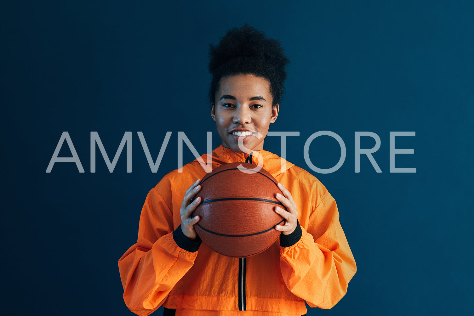 Young woman in orange fitness attire posing over a blue background. Female with basketball looking at camera in studio.