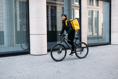 Smiling courier girl riding a bicycle wearing delivery thermal backpack looking for a customer address