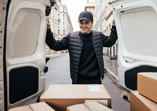 Smiling man in uniform and cap looking at cardboard boxes while standing at van trunk