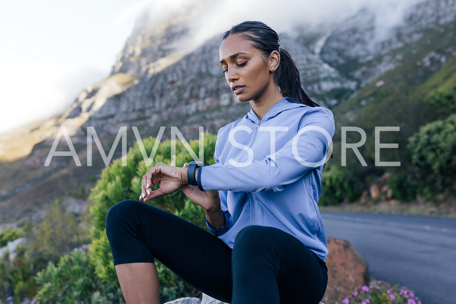 Young sportswoman checking pulse while sitting on a rock in a natural park. Female looking at smartwatch while sitting outdoors in a natural park. 