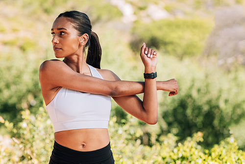 Slim and muscular female athlete flexing her hands and looking away. Young woman exercising outdoors on sunny day.