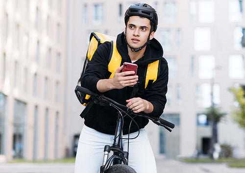 Portrait of young courier leaning on handlebar of a bike holding mobile phone looking for an address for delivery