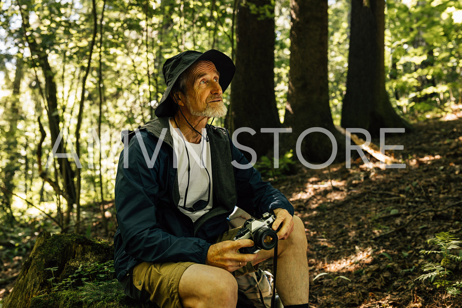 Senior male sitting in the forest and holding a film camera. Mature male in a hat and hiking clothes taking a break during a forest walk.