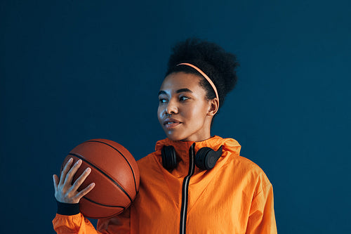 Young female with basketball and wireless headphones posing in studio. Confident sportswoman in orange fitness attire over blue background looking away.