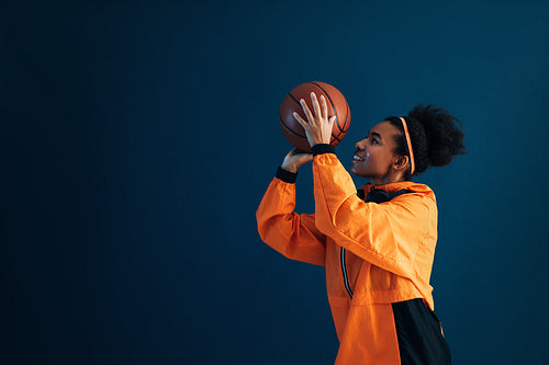 Basketball player in orange sportswear throwing a basketball over a blue background. Side view of professional basketball player practicing over blue backdrop.