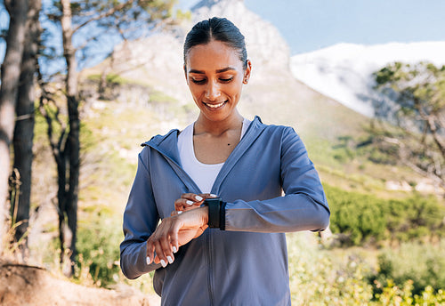 Cheerful woman checking her smartwatches after a workout in a natural park. Smiling slim female checking pulse on a fitness tracker.