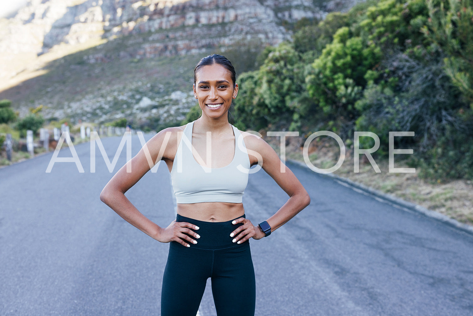 Smiling confident sportswoman standing on abandoned road with hands on hips and looking at camera 