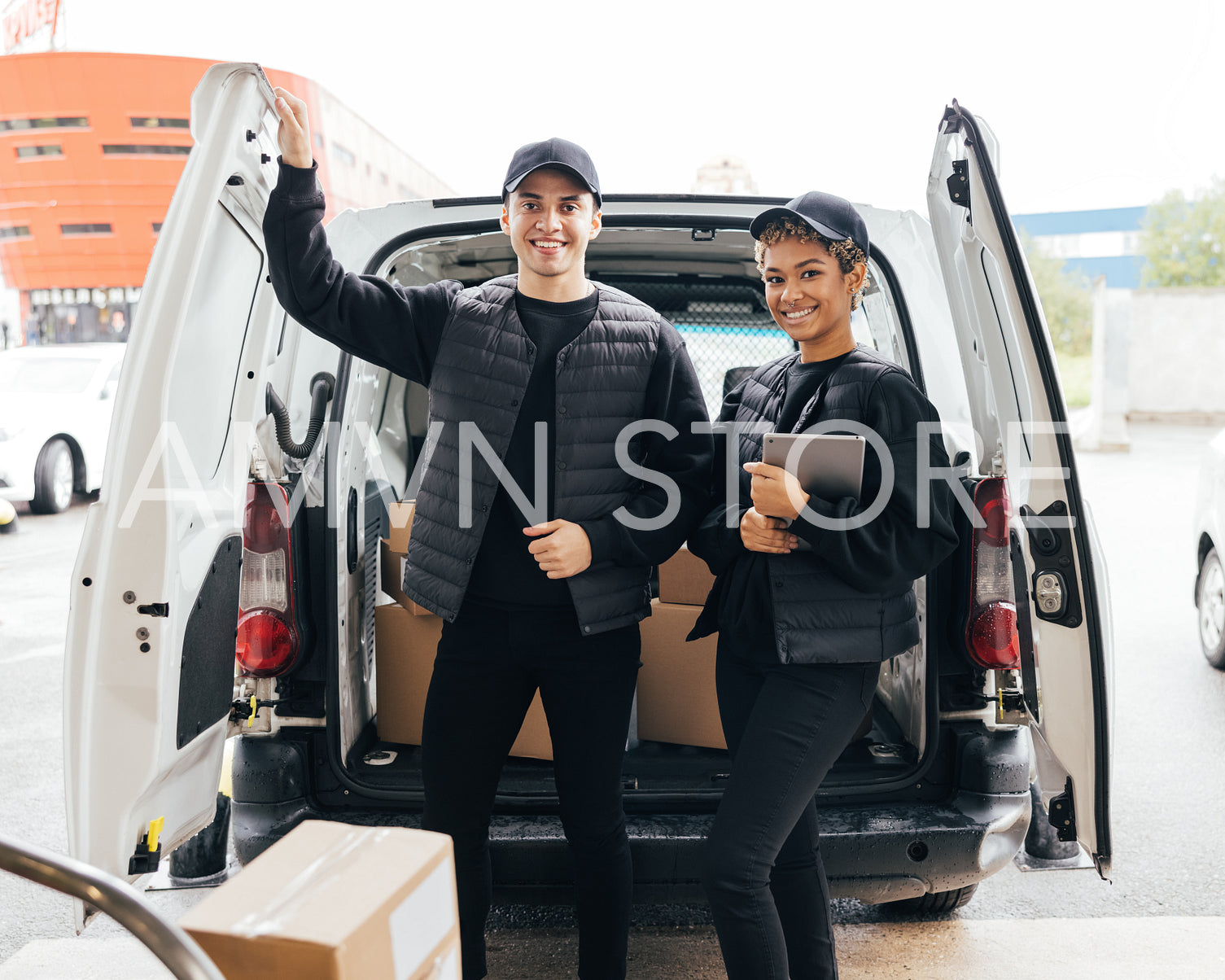 Portrait of two young couriers in uniform standing at van trunk and looking at camera