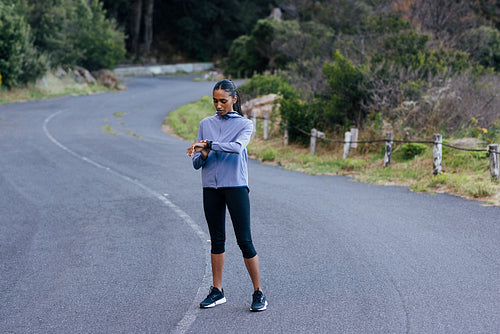 Full length of young slim female checking pulse during exercise while standing in the abandoned road. Female running taking a break checking pulse on smartwatches.