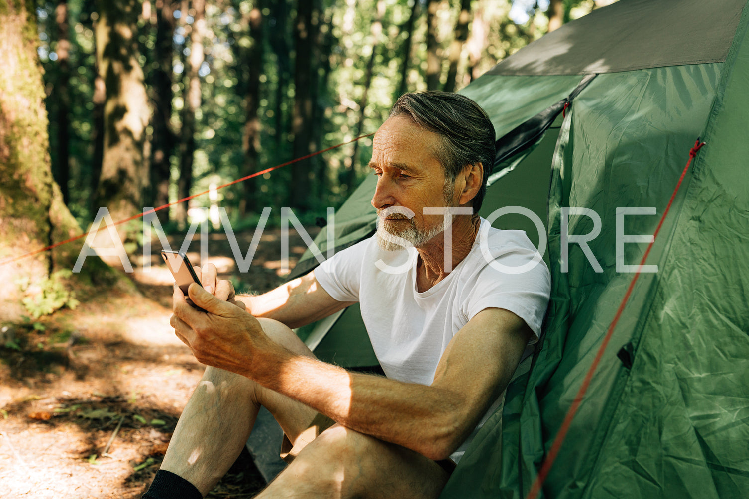 Mature male sitting in a tent and using a smartphone. Senior man with a smartphone in the forest.