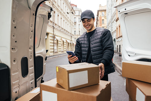 Smiling courier in uniform standing at car trunk checking mobile