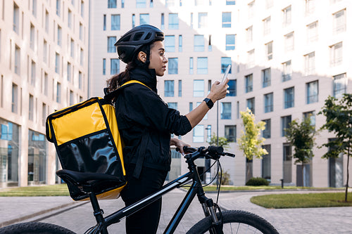 Side view of delivery woman with backpack looking at smartphone while standing in the city against apartment building