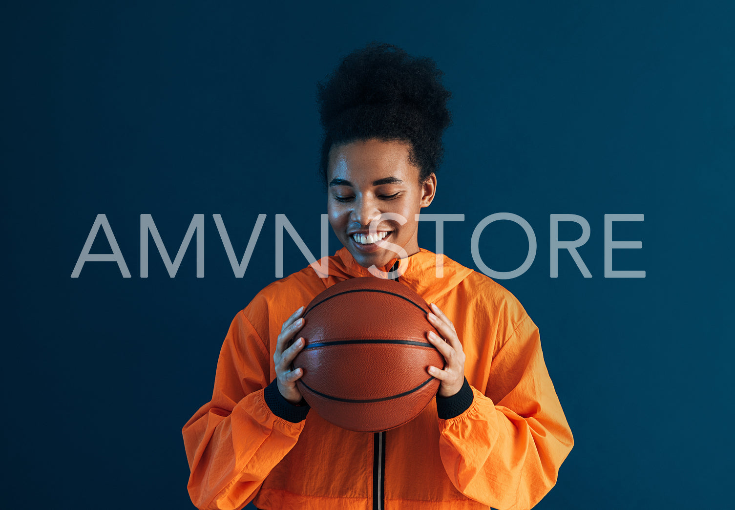 Positive female in orange sportswear holding a basketball. Young woman with closed eyes over blue backdrop holding a basketball.