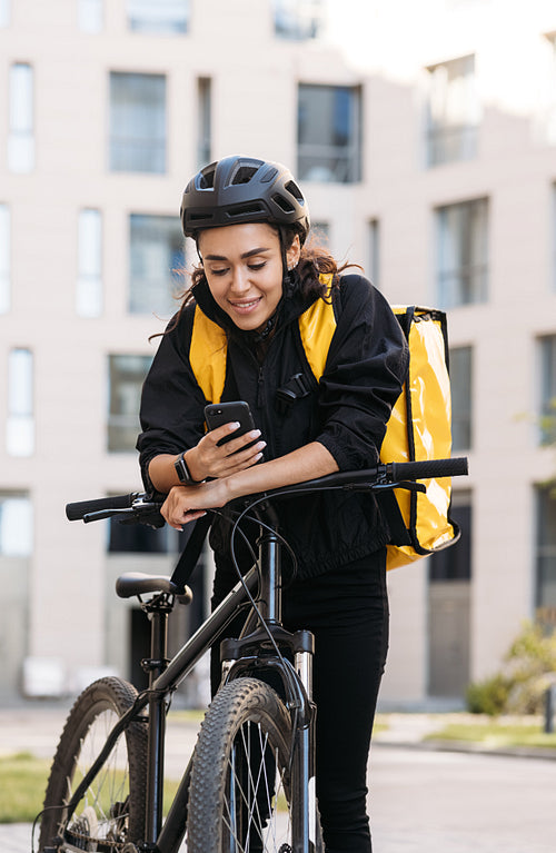 Smiling woman messenger with bicycle and thermal backpack holding a smartphone in the city