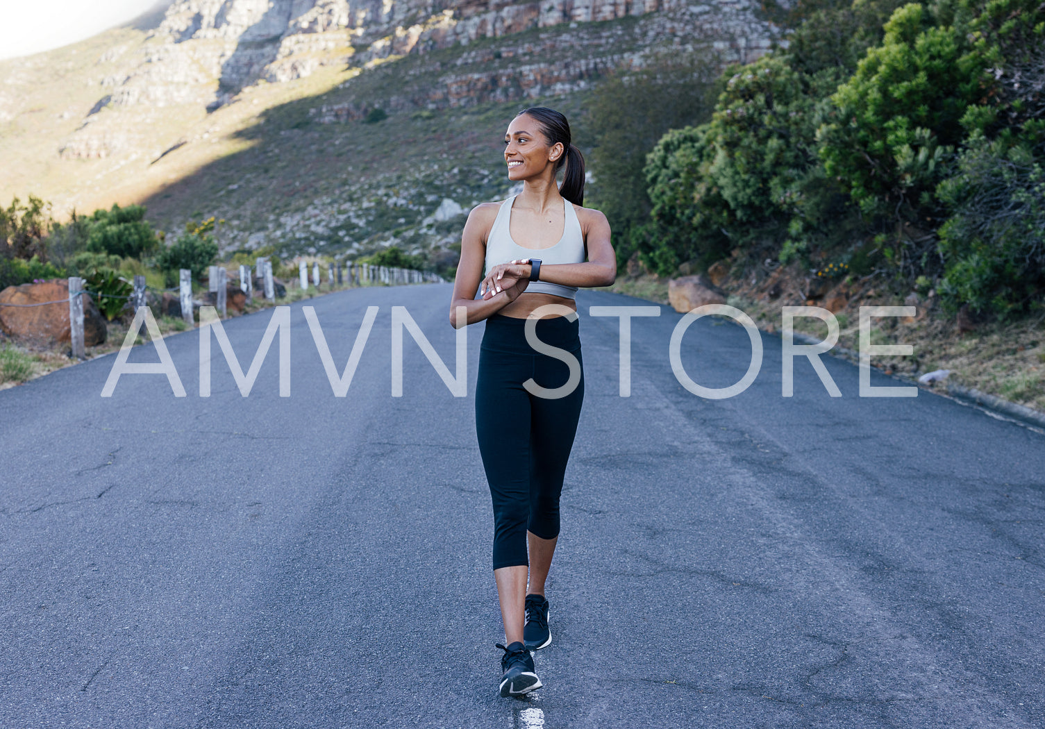 Slim smiling woman walking in the middle of the abandoned road checking her smartwatches. Full length of a young cheerful sports female looking away while walking on the road against a mountain.