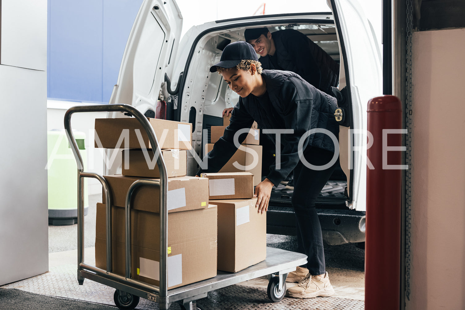 Female courier loads cardboard boxes from a cart into a van whil