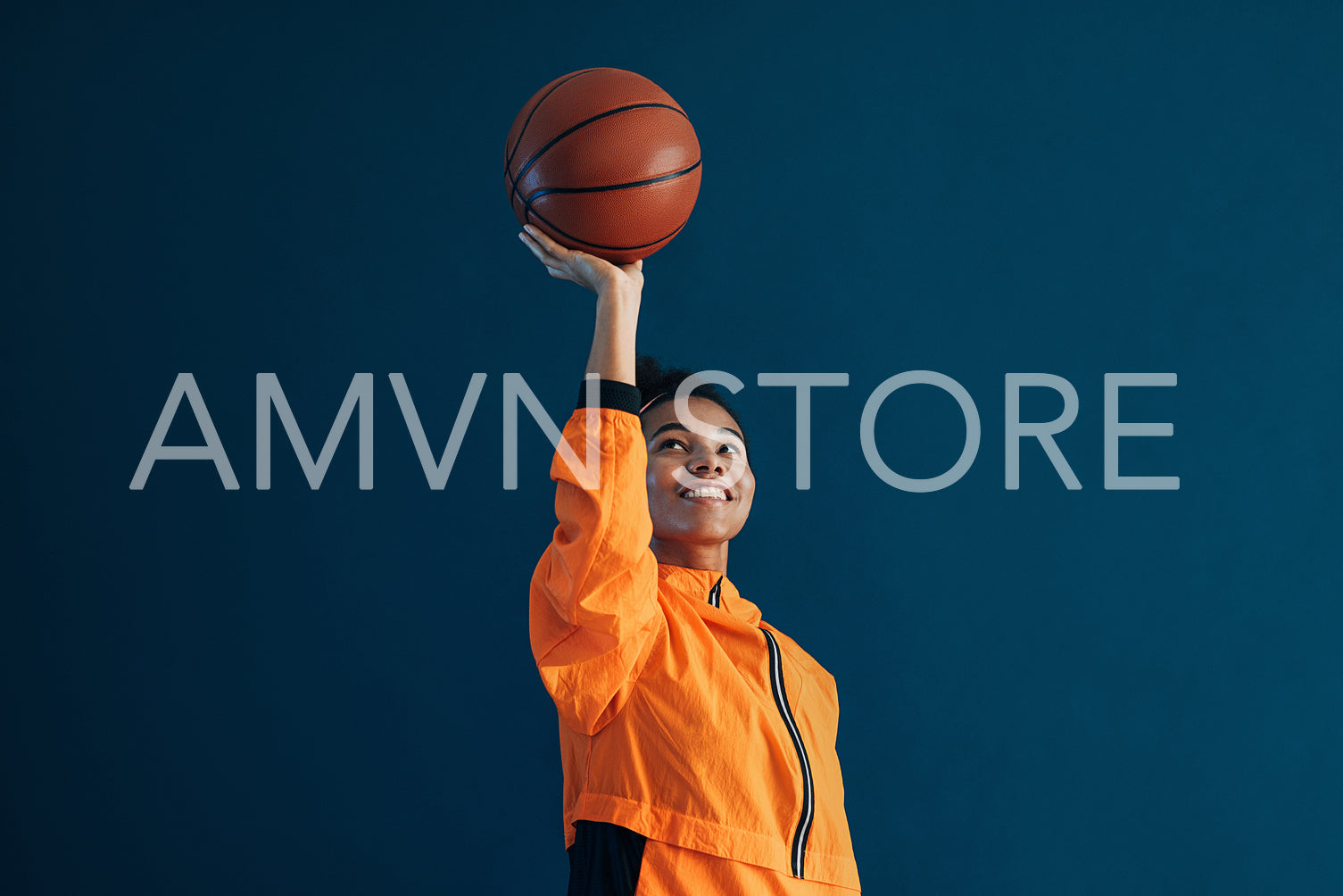 Cheerful female with curly hair holding a basketball over blue backdrop. Smiling professional basketball player holding a basketball in an outstretched hand.