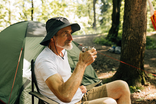 Side view of senior male with the cup sitting in a chair against the tent in forest