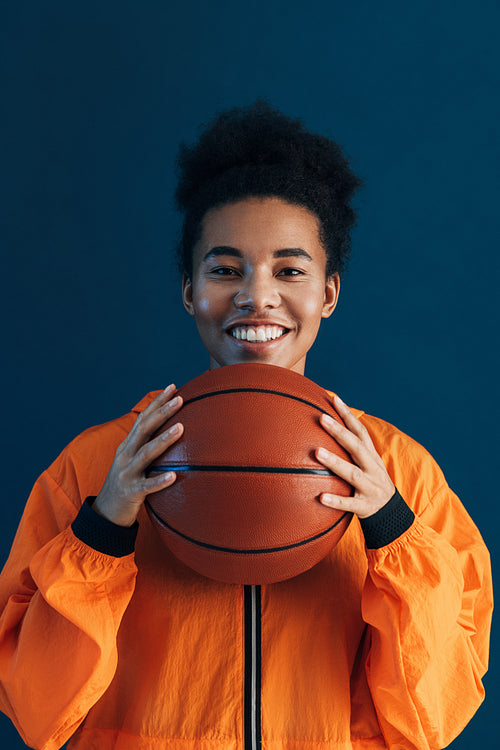 Portrait of a young smiling basketball player. Cheerful female in orange sportswear holding basketball over blue backdrop.
