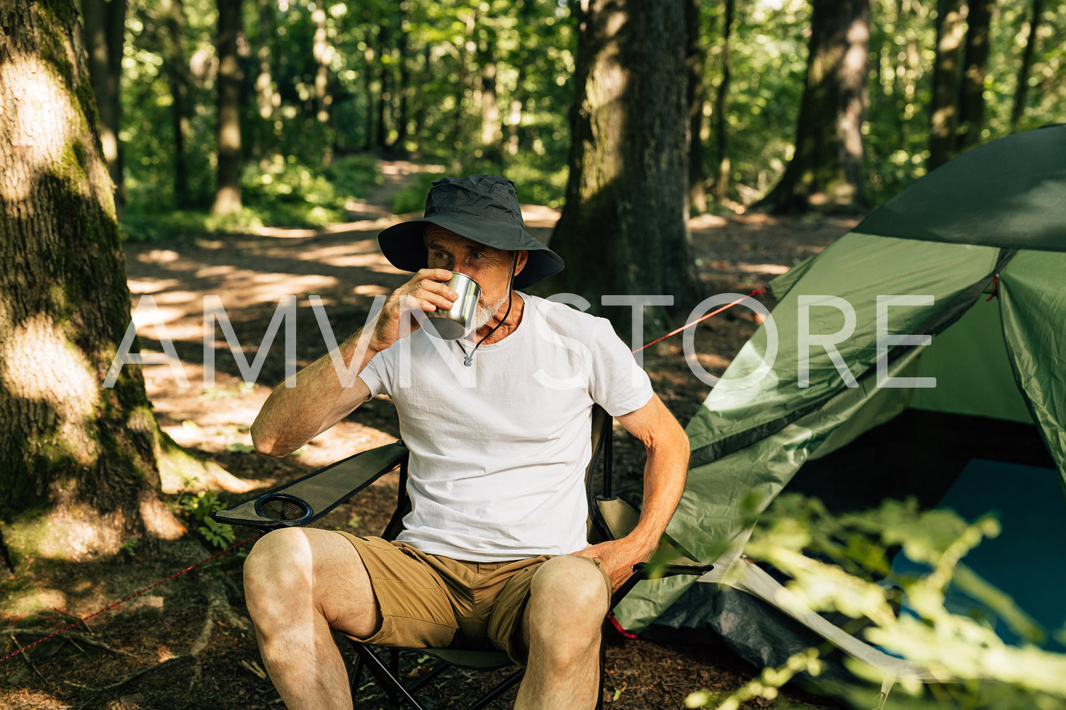 Senior tourist drinks coffee while sitting on chair in the forest near his tent