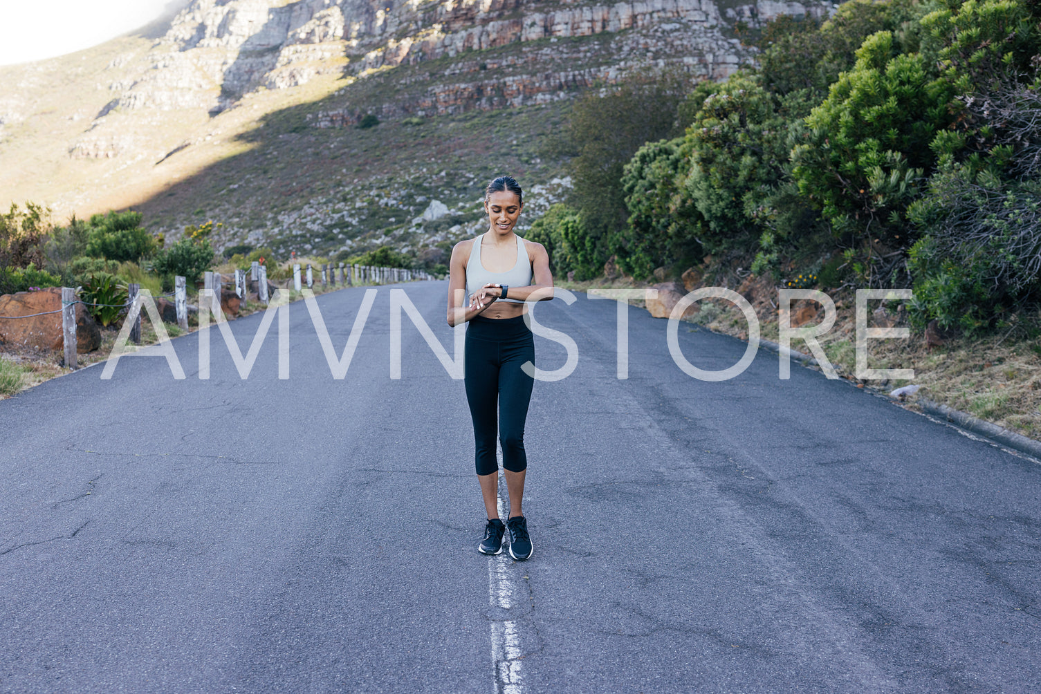 Full length of a smiling female walking on an abandoned road and checking her pulse. Cheerful woman using smartwatches while doing morning walk.