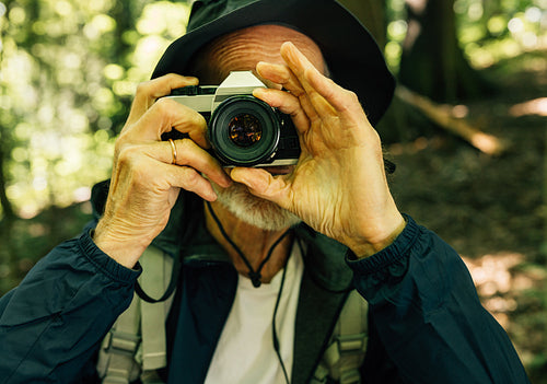 Close-up of a senior male in a hat taking pictures on a film camera. Mature man with a film camera in forest.