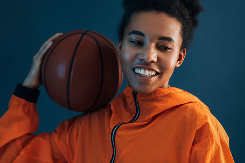 Close-up of young professional female basketball player over blue backdrop. Cheerful woman in orange sportswear with basketball on her shoulder.