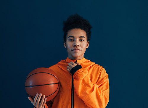 Young female adjusting her orange sportswear and looking at the camera over a blue backdrop. Professional female basketball player with the ball in the studio.