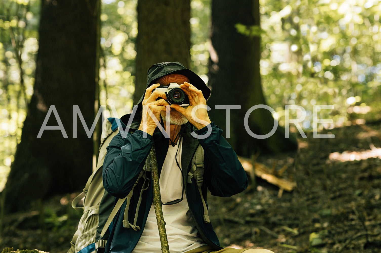 Senior tourist in hat taking photographs while sitting in a forest during a hike