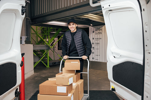 Smiling courier in uniform standing in warehouse with cart prepa