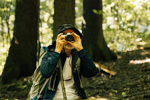 Senior tourist in hat taking photographs while sitting in a forest during a hike