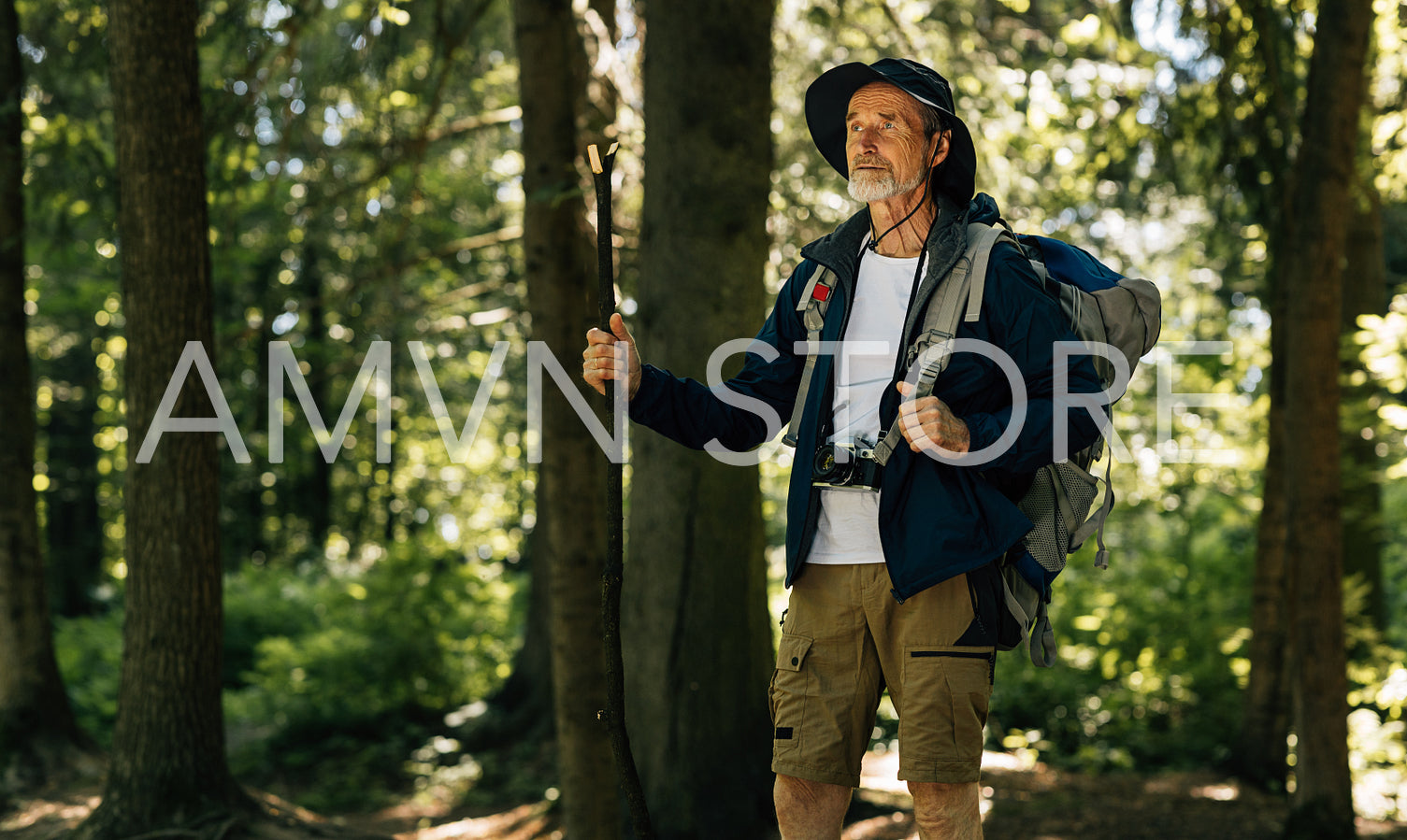 Senior man wearing hiking clothes and accessories standing in forest. Mature tourist with wooden stick enjoying hike.