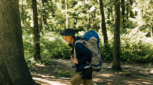 Side view of a senior male walking with touristic backpack in a forest during a sunny day