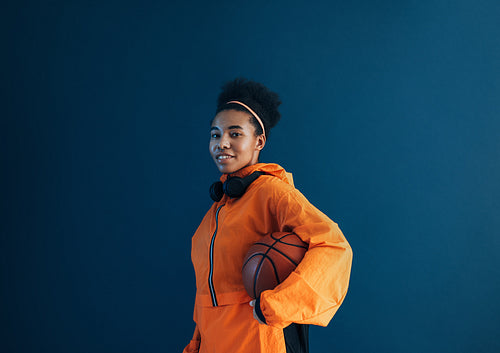 Young sportswoman standing with basketball in studio and looking at camera. Female in orange sportswear over blue background.